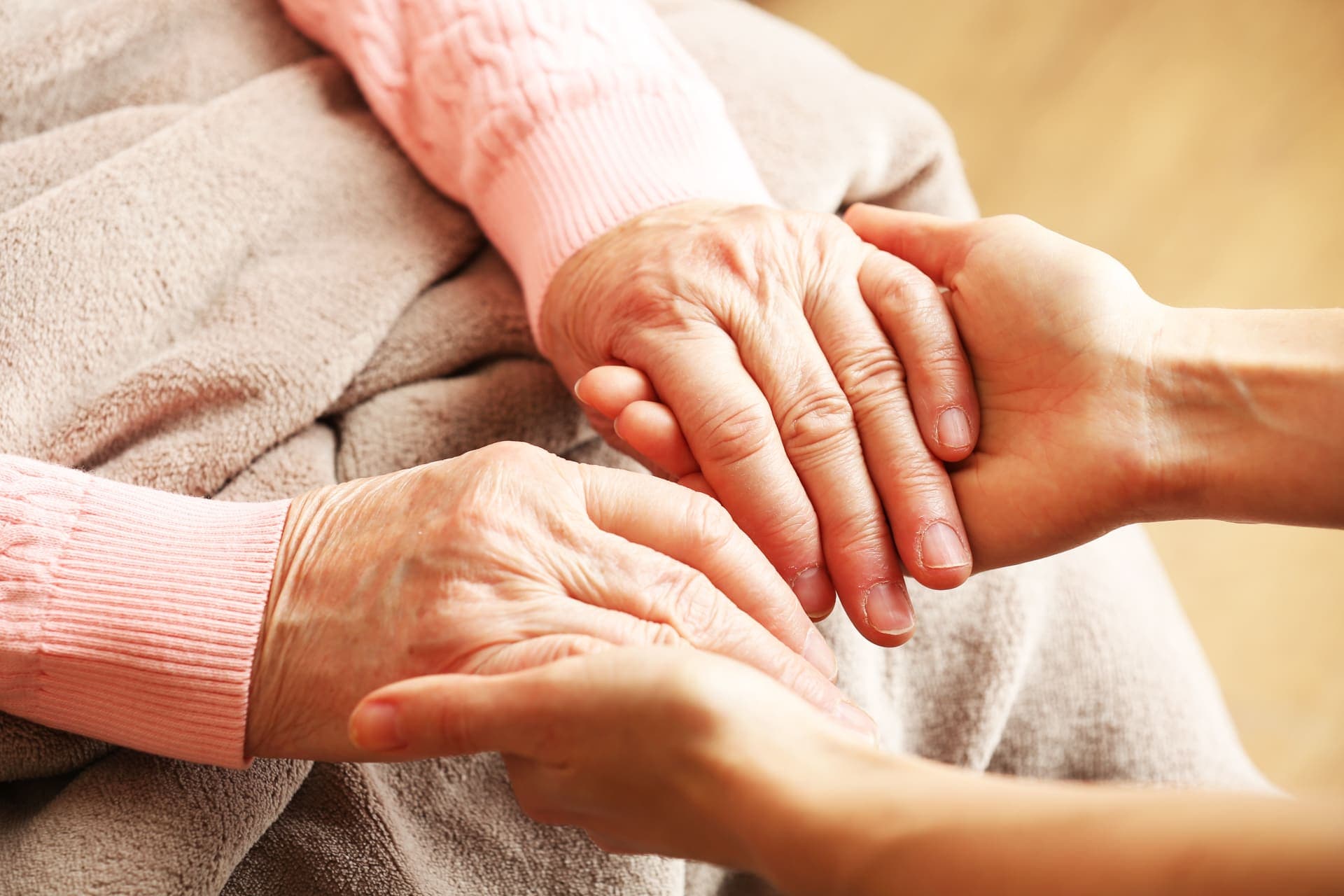 A younger person's hands cradling the hands of an older woman in a soft pink cardigan