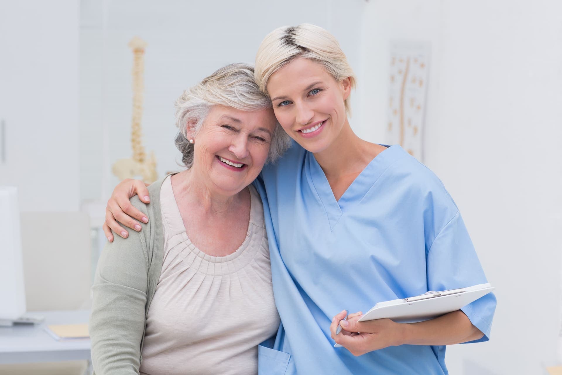 An older woman and a nurse in blue scrubs standing arm in arm, both smiling at the camera